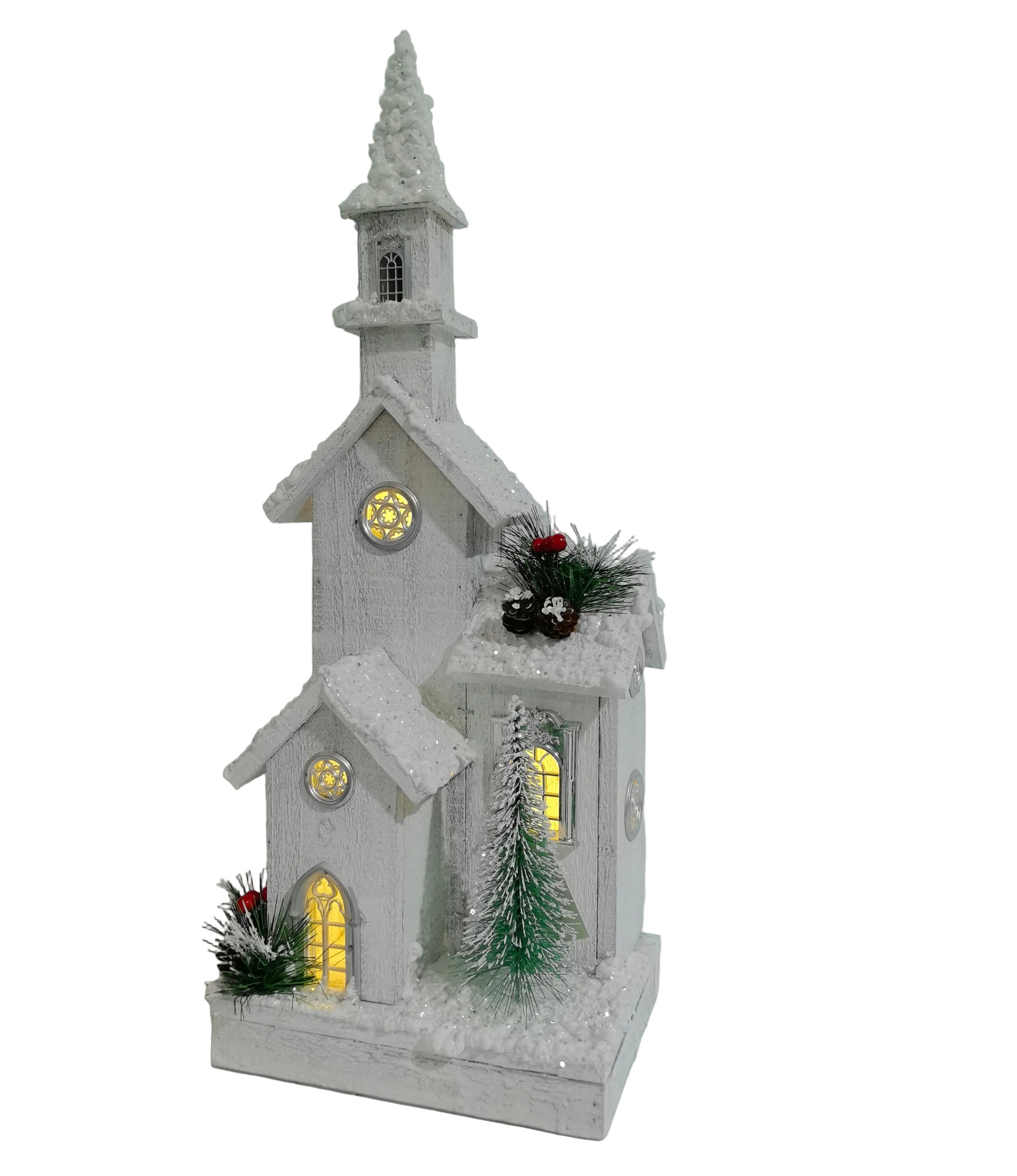 A white Christmas church decoration with a snowy textured finish, glowing yellow windows, green pine branches, and red berries.