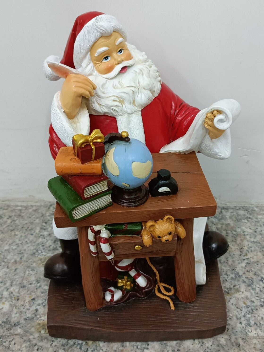 A resin figurine of Santa Claus sitting at a desk with a world globe, books, a teddy bear, and a Christmas stocking.